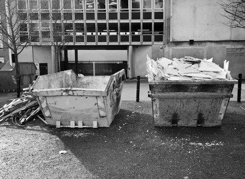 Two large industrial skip bins positioned side by side on a paved area in front of a modern building with a glass frontage and concrete exterior walls. The left skip contains large, irregularly shaped concrete debris with rough, chipped surfaces and a light grey coloration. The right skip is filled with multiple white paper or cardboard sheets haphazardly piled, some crumpled and others flat, with visible edges and creases. Both skips are metal with signs of weathering; the left shows signs of concrete residue and rust, while the right has darker patches and surface wear. The surrounding ground is scattered with small debris, and in the background, there is a view of an urban pathway with metal bollards and a metal fence, indicating an environment typical of commercial or office clearance waste removal in Barkingside. The overall scene appears to be part of a rubbish collection or clearance process managed by Office Clearance Barkingside, serving the local area postcodes within the Barkingside town area.