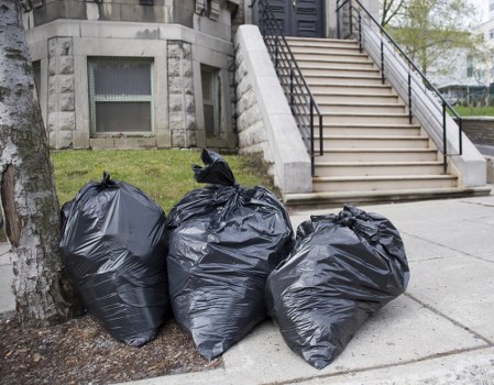 Three large black rubbish bags made of thick plastic with a slightly glossy surface are stacked against a tree to the left of a concrete sidewalk. The bags are full and tied at the top, with some slight creases and folds visible in the material. Behind the bags, there is a small area of well-maintained grass and the base of a modern building with light grey stone walls and two small, rectangular windows with frosted glass. To the right, a wide set of outdoor stairs with metal handrails on both sides leads up to an entrance elevated from the pavement. The scene is set outdoors in an urban environment, possibly in Barkingside, near a residential or commercial property. This image represents typical rubbish collection for waste clearance services, such as those provided by Office Clearance Barkingside, focusing on outdoor waste removal with a clean, professional appearance.