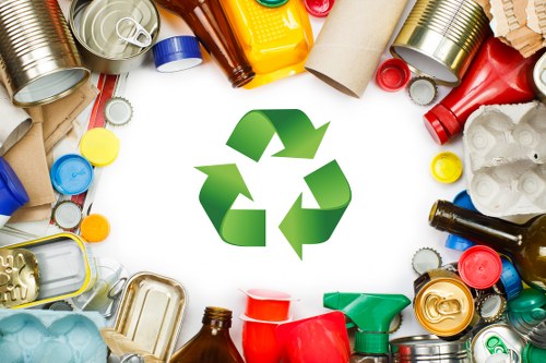 A collection of assorted waste items arranged around a central green recycling symbol. The waste includes various discarded containers such as aluminum cans, plastic bottles, and glass bottles, along with some cardboard tubes, lids, and foil packaging. The objects are scattered across a plain white surface, with some items overlapping each other. The aluminium cans are metallic with shiny finishes, while the plastic bottles are in different colours, including red, yellow, and brown, some with labels still attached. The cardboard pieces are light brown with visible corrugation. The overall scene is clean, organized, and visually highlights typical household or office waste that Office Clearance Barkingside might remove as part of rubbish clearance services in the Barkingside area, close to postcode IG6. The lighting is neutral, emphasizing the textures and colours of the waste materials without shadows or reflections.