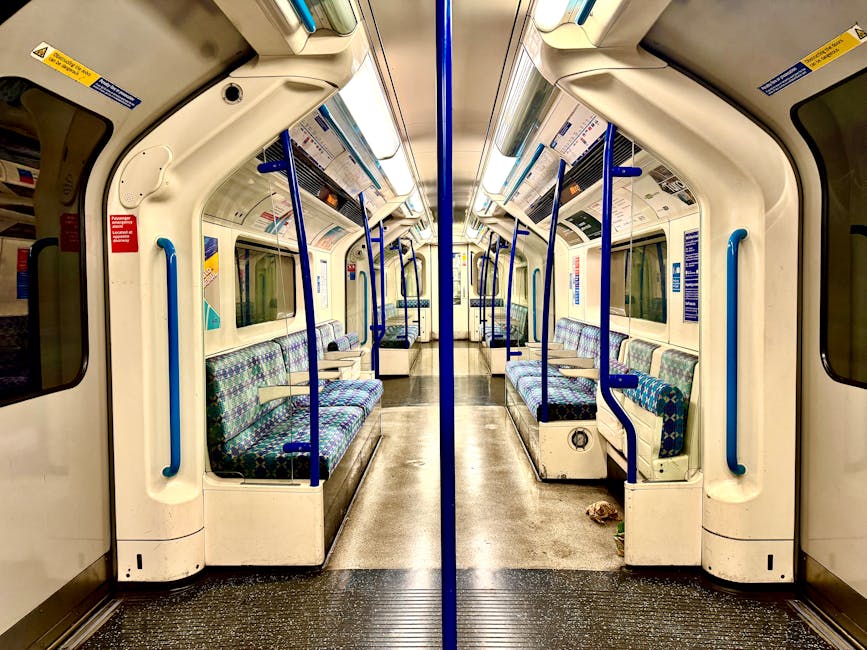 A section of a London underground train station platform with a partially visible modern train showing white, orange, and blue exterior livery parked alongside the platform. The platform has textured 