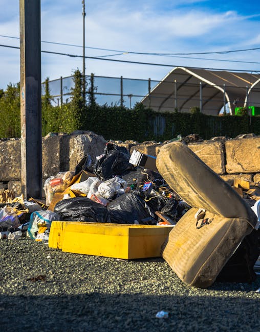 A ground-level view of an outdoor driveway in a residential area showing a collection of various waste materials ready for removal. Prominently in the foreground, there are multiple large black and bl